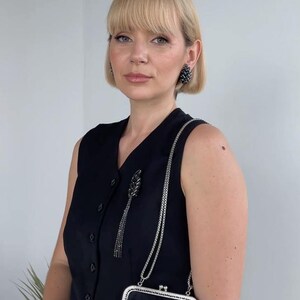 May include: A woman wearing a black sleeveless dress with a silver chain strap purse. The purse has a black velvet exterior with a floral design and rhinestones. She is also wearing black rhinestone earrings.