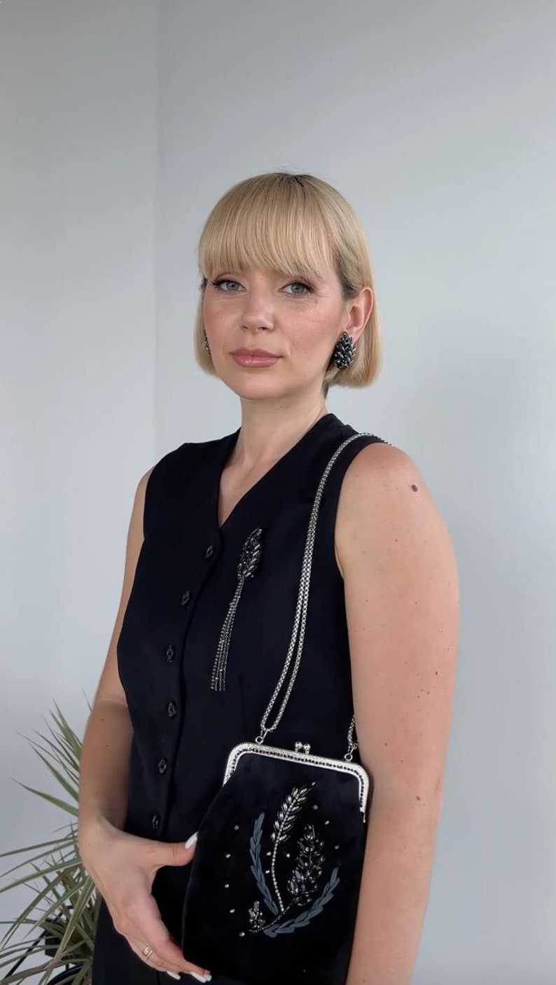 May include: A woman wearing a black sleeveless dress with a silver chain strap purse. The purse has a black velvet exterior with a floral design and rhinestones. She is also wearing black rhinestone earrings.