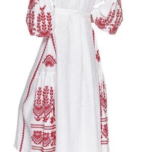 May include: White linen dress with red embroidered floral patterns. The dress has a cinched waist and long sleeves with a gathered puff at the shoulder.