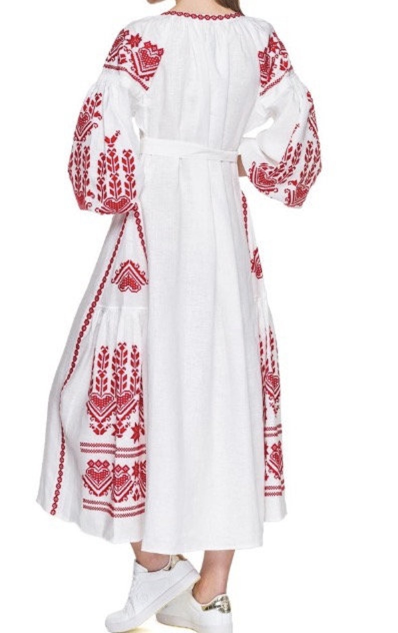 May include: White linen dress with red embroidered floral patterns. The dress has a cinched waist and long sleeves with a gathered puff at the shoulder.