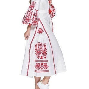 A white linen dress with red embroidered floral patterns. The dress has a long, flowing skirt and a fitted bodice. The sleeves are long and puffy. The dress is belted at the waist and has a v-neckline.