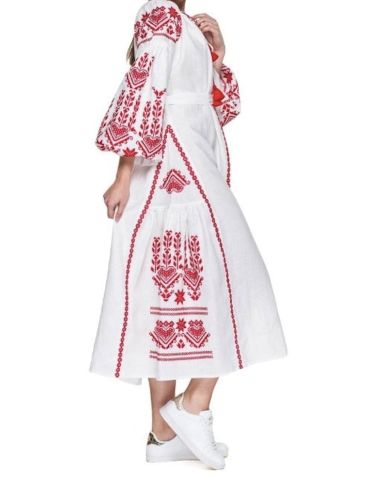 A white linen dress with red embroidered floral patterns. The dress has a long, flowing skirt and a fitted bodice. The sleeves are long and puffy. The dress is belted at the waist and has a v-neckline.