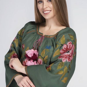 Puede incluir: Una blusa de lino verde con detalles florales bordados y borlas rosas y burdeos. La blusa tiene un escote redondo y mangas largas.