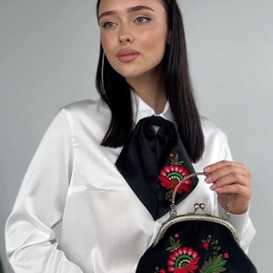 Pode incluir: Uma mulher vestindo uma blusa de cetim branco com uma gravata de la&ccedil;o de cetim preto e cal&ccedil;as pretas. Ela est&aacute; segurando uma bolsa de embreagem de veludo preto com um design de bordado floral em vermelho, verde e dourado.