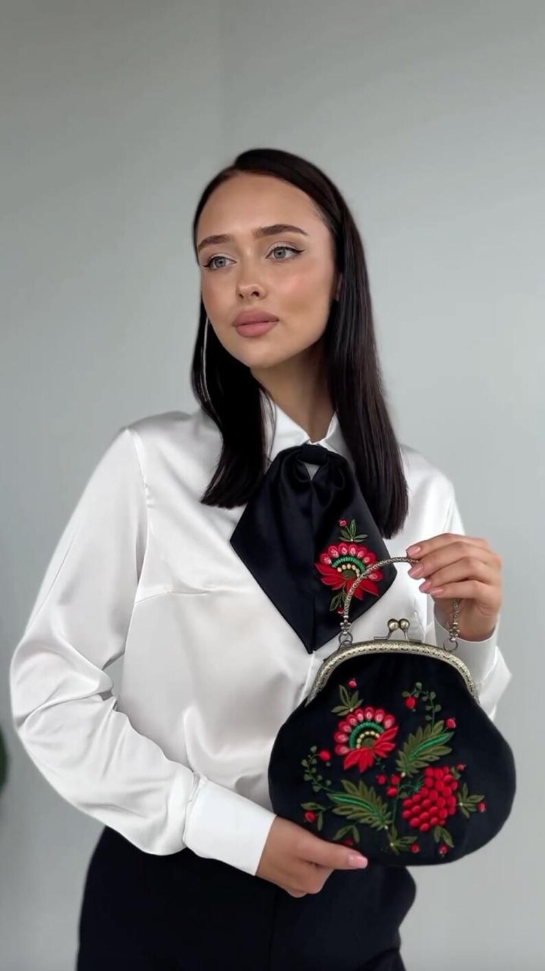Pode incluir: Uma mulher vestindo uma blusa de cetim branco com uma gravata de la&ccedil;o de cetim preto e cal&ccedil;as pretas. Ela est&aacute; segurando uma bolsa de embreagem de veludo preto com um design de bordado floral em vermelho, verde e dourado.