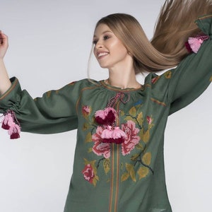 Puede incluir: Una blusa de lino verde con flores bordadas en rosa y rojo. La blusa tiene mangas largas con borlas rosas y rojas en los pu&ntilde;os.
