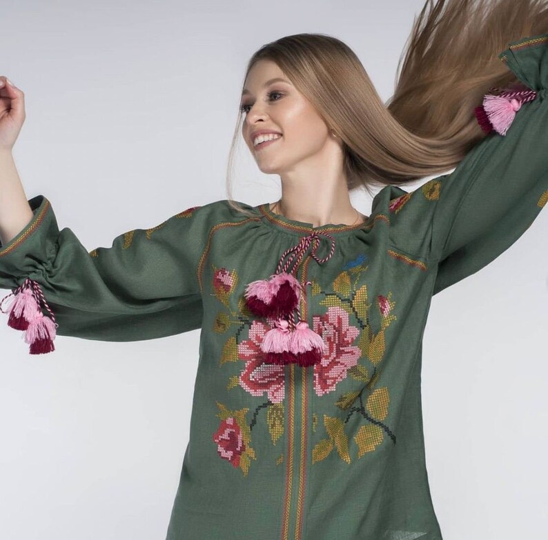 Puede incluir: Una blusa de lino verde con flores bordadas en rosa y rojo. La blusa tiene mangas largas con borlas rosas y rojas en los pu&ntilde;os.