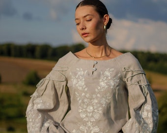 May include: A woman wearing a light gray linen blouse with white embroidery. The blouse has a relaxed fit and long sleeves with a gathered detail at the cuffs. The woman is standing in a field with green grass and a blue sky in the background.