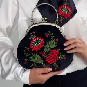Pode incluir: Uma bolsa de veludo preto com um design de bordado floral com flores vermelhas e verdes. A bolsa tem um fecho prateado e uma al&ccedil;a de corrente prateada.
