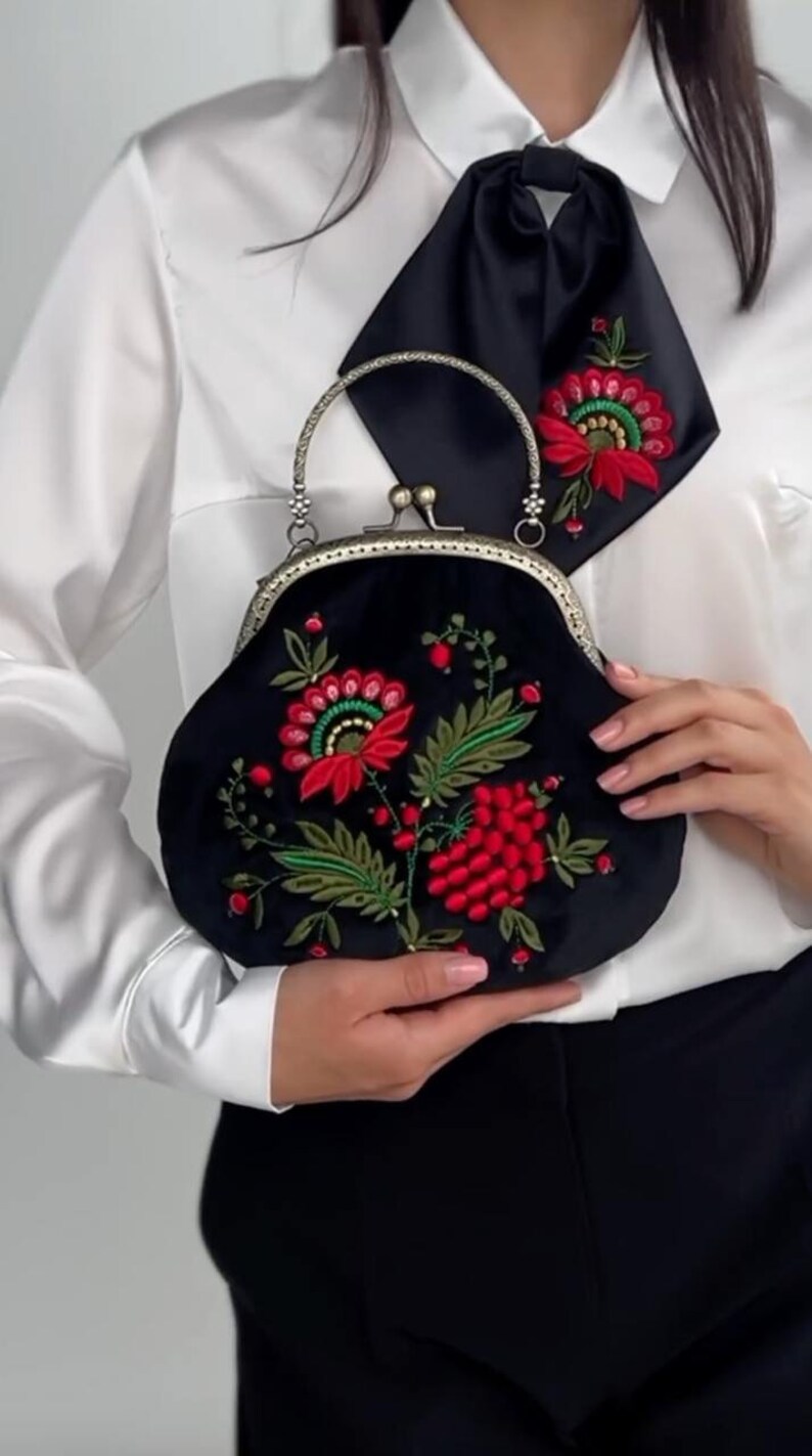 Pode incluir: Uma bolsa de veludo preto com um design de bordado floral com flores vermelhas e verdes. A bolsa tem um fecho prateado e uma al&ccedil;a de corrente prateada.