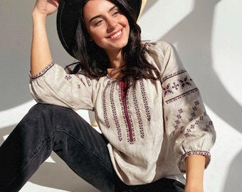Puede incluir: Una mujer lleva una blusa de lino beige con bordados marrones y pantalones vaqueros negros. Lleva un sombrero negro de ala ancha y tiene el pelo recogido hacia atrás.