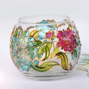 May include: A clear glass bowl featuring hand-painted floral designs in vibrant shades of turquoise, magenta, and green. The artwork includes detailed petals and leaves outlined in gold, with small raised dots accenting the floral patterns.