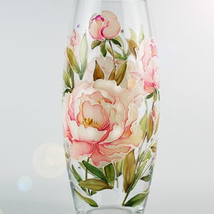 Puede incluir: Un jarrón de vidrio transparente con un diseño floral pintado. El jarrón presenta grandes flores de peonía de color rosa claro con centros amarillos y hojas verdes. Las flores están pintadas en el exterior del jarrón.