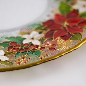 May include: A clear glass plate with a gold rim, decorated with hand-painted Christmas floral designs. The design includes red poinsettias, white flowers, green leaves, and red berries. The plate has a scalloped edge.