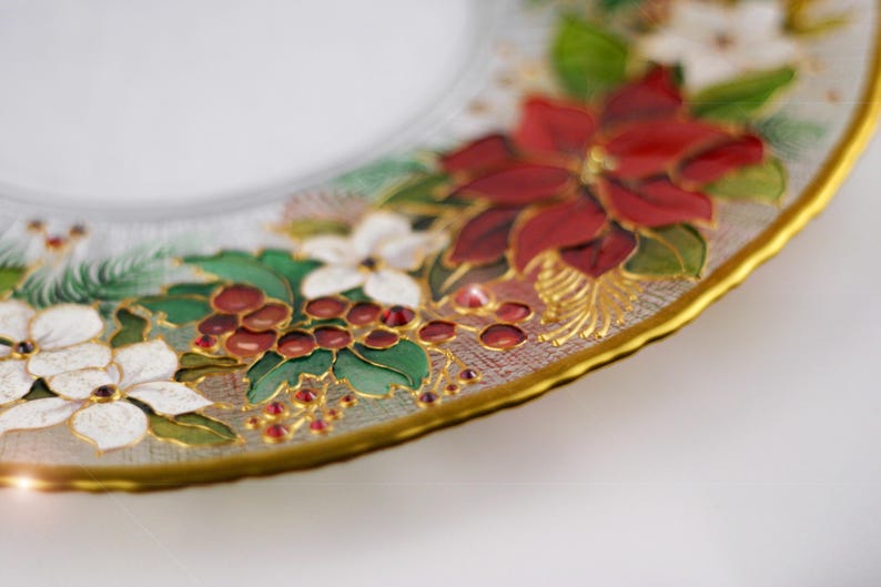 May include: A clear glass plate with a gold rim, decorated with hand-painted Christmas floral designs. The design includes red poinsettias, white flowers, green leaves, and red berries. The plate has a scalloped edge.