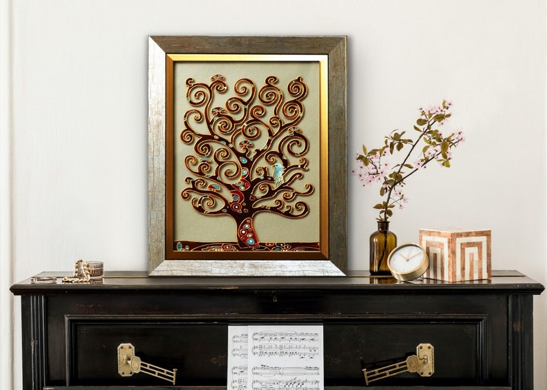 May include: A framed artwork featuring a stylized tree with swirling branches in shades of brown, gold, and blue. The tree is set against a beige background and framed in a silver and gold frame.