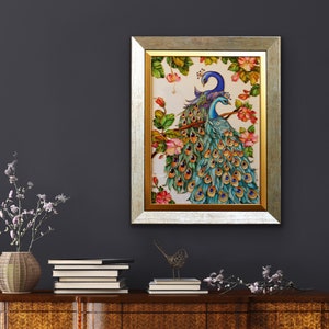 May include: A framed artwork featuring two peacocks perched on a branch with pink flowers. The peacocks have blue and teal plumage. The frame is gold and silver. The artwork is displayed on a dark gray wall.