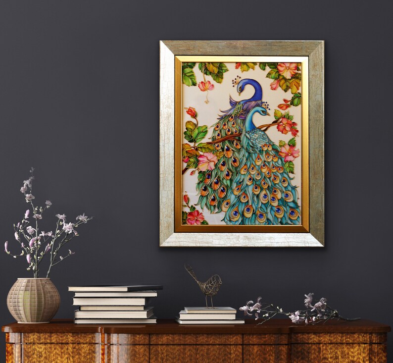 May include: A framed artwork featuring two peacocks perched on a branch with pink flowers. The peacocks have blue and teal plumage. The frame is gold and silver. The artwork is displayed on a dark gray wall.