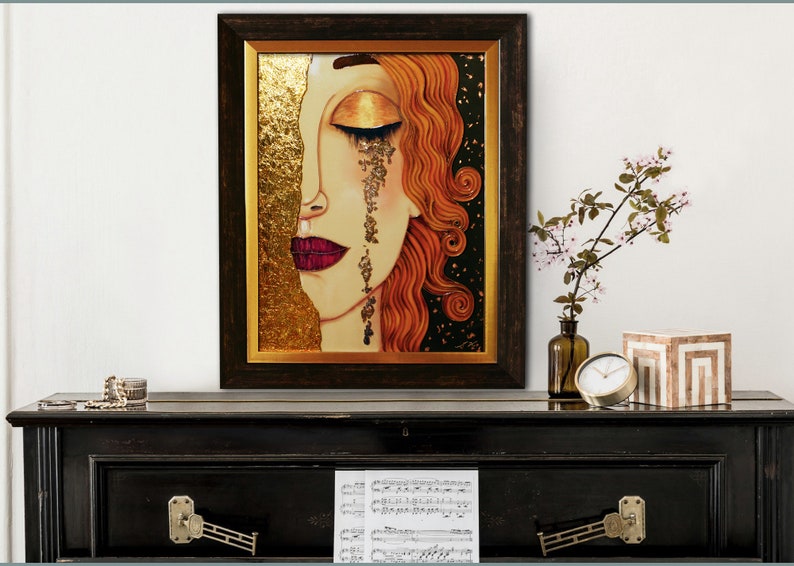 Puede incluir: Una pintura enmarcada de la cara de una mujer con detalles de pan de oro. Sus ojos est&aacute;n cerrados y est&aacute; llorando l&aacute;grimas de oro. El fondo es negro con motas doradas.