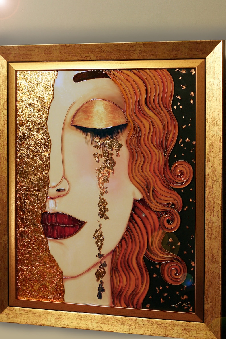 Puede incluir: Una pintura enmarcada de la cara de una mujer con los ojos cerrados y una sola l&aacute;grima hecha de pan de oro. El cabello de la mujer es rojo y fluido, y el fondo es negro con motas doradas.