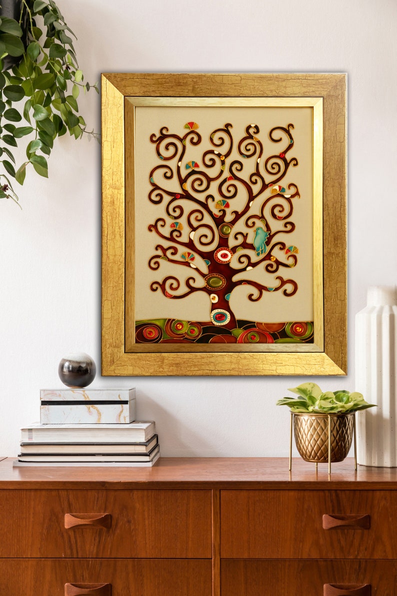 May include: A framed artwork featuring a stylized tree with a brown trunk and branches, decorated with colorful circles and swirls. The tree is set against a beige background and framed in a gold frame.
