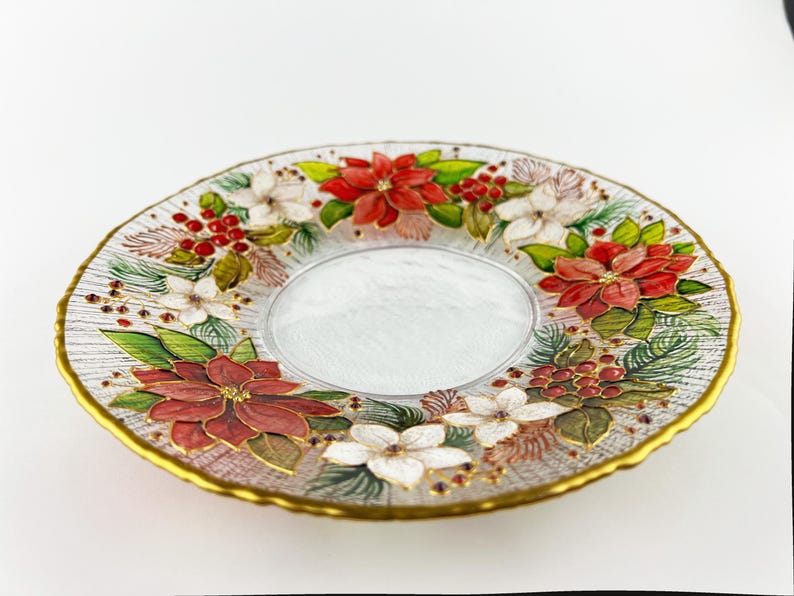 May include: A clear glass plate with a gold rim, decorated with red poinsettias, white flowers, green leaves, and red berries. The center of the plate is a clear, oval shape.