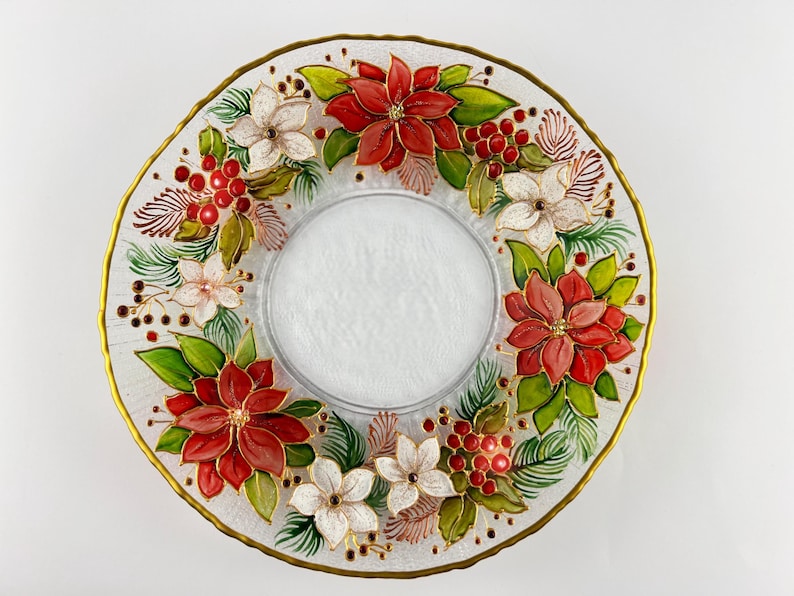 May include: A clear glass decorative plate with a gold rim, featuring a poinsettia and berry design. The plate has a central recessed area and is adorned with red, white, and green floral elements. A festive holiday accent.