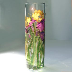 Può includere: Un vaso cilindrico in vetro trasparente decorato con fiori di iris dipinti a mano in giallo e viola con steli verdi. Il design presenta contorni color oro e piccole decorazioni trasparenti applicate sulla superficie del vetro.