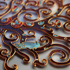May include: A decorative glass art piece featuring a stylized tree with intricate gold and brown swirls. A small blue bird sits on a branch. The piece is painted with various colors, including blue, red, and gold.