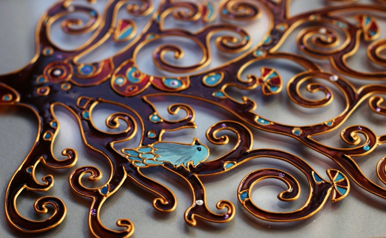 May include: A decorative glass art piece featuring a stylized tree with intricate gold and brown swirls. A small blue bird sits on a branch. The piece is painted with various colors, including blue, red, and gold.