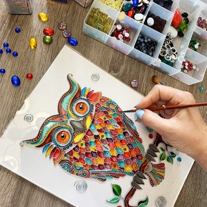 May include: A colorful owl painting on a white canvas with a hand holding a paintbrush and a container of colorful rhinestones. The owl is painted in a variety of colors, including red, orange, yellow, green, blue, and purple. The owl is surrounded by a variety of swirls and other decorative elements.
