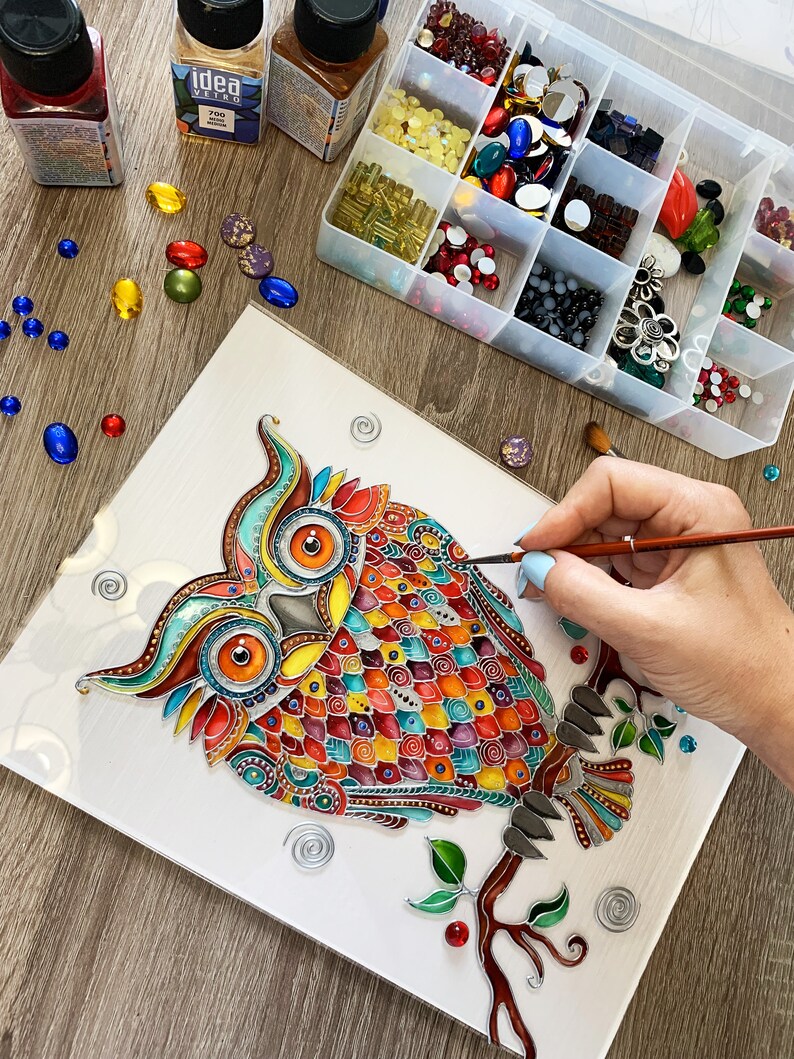 May include: A colorful owl painting on a white canvas with a hand holding a paintbrush and a container of colorful rhinestones. The owl is painted in a variety of colors, including red, orange, yellow, green, blue, and purple. The owl is surrounded by a variety of swirls and other decorative elements.