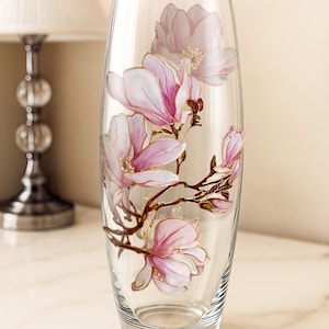 Peut inclure: Un vase en verre transparent avec un motif floral peint. Le vase pr&eacute;sente des fleurs de magnolia roses avec des accents dor&eacute;s sur des branches brunes. Le vase est de forme ovale et repose sur une surface en marbre.