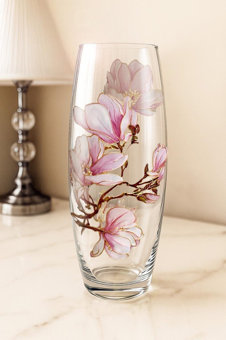 Peut inclure: Un vase en verre transparent avec un motif floral peint. Le vase pr&eacute;sente des fleurs de magnolia roses avec des accents dor&eacute;s sur des branches brunes. Le vase est de forme ovale et repose sur une surface en marbre.