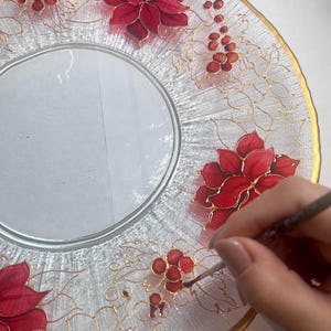 May include: Clear glass plate with a gold rim and intricate hand-painted red poinsettias and berries. Delicate gold vines connect the floral elements. The plate is being painted with a fine brush, showcasing the artistry.