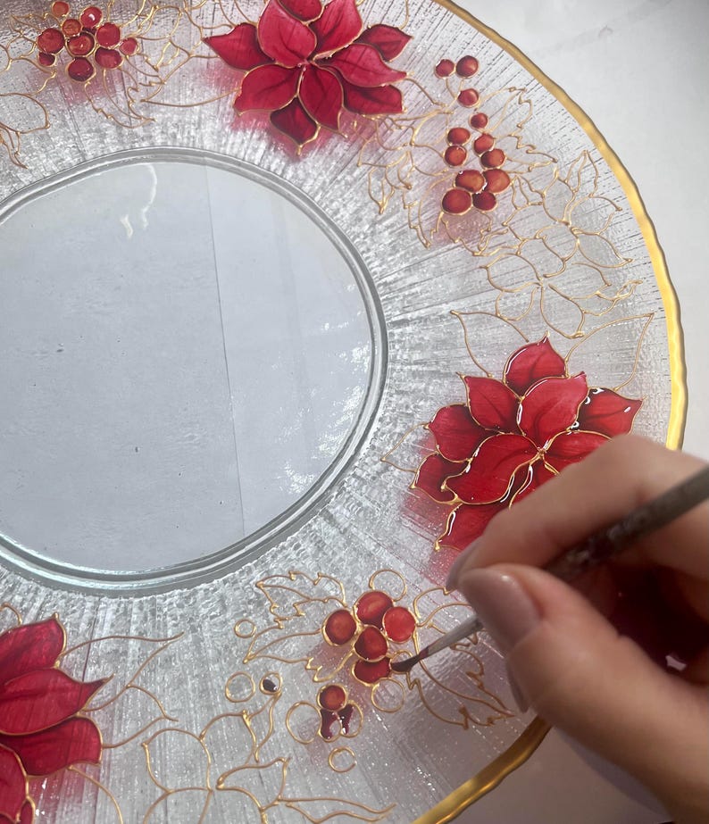 May include: Clear glass plate with a gold rim and intricate hand-painted red poinsettias and berries. Delicate gold vines connect the floral elements. The plate is being painted with a fine brush, showcasing the artistry.