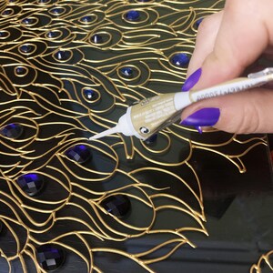 May include: A close-up of a peacock feather design with gold-colored outlines, dark blue accents, and blue gemstones. A tube of adhesive is being used to apply glue to the design. The design is on a black surface.