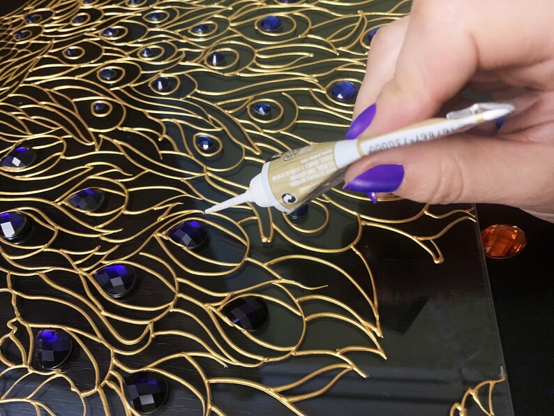 May include: A close-up of a peacock feather design with gold-colored outlines, dark blue accents, and blue gemstones. A tube of adhesive is being used to apply glue to the design. The design is on a black surface.