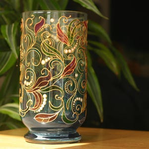 Può includere: Un vaso di vetro blu con un design dipinto a mano di motivi floreali dorati, verdi e rossi. Il vaso ha una base rotonda e un corpo cilindrico.