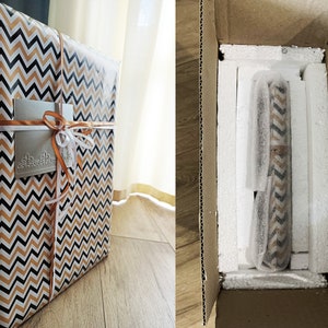 May include: A gift box wrapped in black and orange chevron patterned paper with a white ribbon and a silver gift tag. The box is sitting on a wooden surface. The box is being placed into a larger cardboard box with white styrofoam packing peanuts.