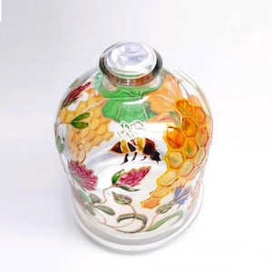 May include: A clear glass cloche with a decorative bee and honeycomb design. The dome-shaped cover features hand-painted details of a bee, honeycomb, and floral elements in shades of yellow, orange, green, and pink.