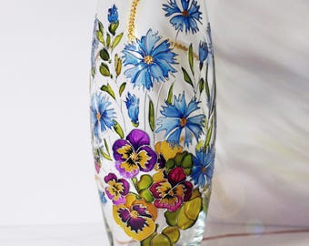 May include: A clear glass vase featuring hand-painted floral designs. The artwork displays vibrant blue cornflowers and multi-colored pansies in shades of purple, yellow, and deep red, accented with green leaves and thin gold-colored stems.