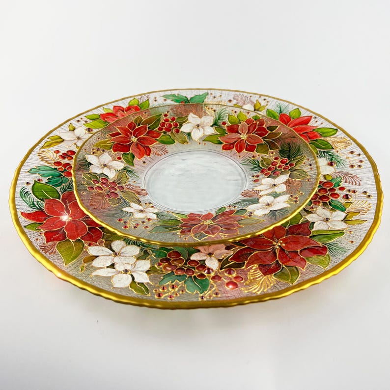 May include: Two-piece glass serving dish set. The larger plate has a gold rim and is decorated with red poinsettias, white flowers, and green leaves. The smaller plate sits on top, featuring a similar floral design.