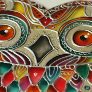 May include: Close-up of a colorful stained glass owl with intricate details. The owl's eyes are large and red with white highlights. The owl's feathers are depicted in shades of red, green, yellow, and blue.