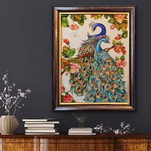May include: A framed artwork featuring two peacocks perched on a branch, surrounded by pink flowers and green leaves. The peacocks have blue and teal plumage with intricate detailing. The frame is dark brown with a gold inner border. The artwork is displayed on a dark wall.