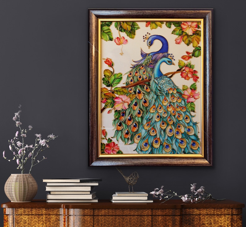 May include: A framed artwork featuring two peacocks perched on a branch, surrounded by pink flowers and green leaves. The peacocks have blue and teal plumage with intricate detailing. The frame is dark brown with a gold inner border. The artwork is displayed on a dark wall.