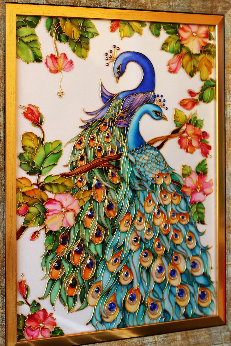 May include: Framed artwork featuring two peacocks with vibrant blue and green plumage, perched on a branch amidst pink flowers and green leaves. The artwork has gold accents and is set against a white background.