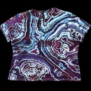 May include: A short-sleeved t-shirt with a tie-dye pattern in shades of purple, blue, and white. The design features a swirling, topographic-like pattern. The shirt is made of a soft material and has a crew neck.