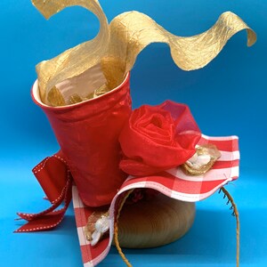 Peut inclure: Un chapeau en vichy rouge et blanc avec une rose rouge, un ruban doré et un accent en feuille d'or. Le chapeau est posé sur une base en bois.