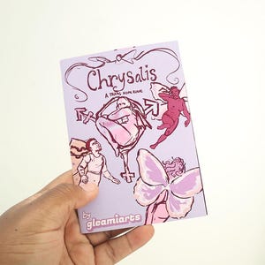 Puede incluir: Un libro de color lavanda titulado "Chrysalis" con ilustraciones en rosa y morado. La portada presenta figuras, flechas y una mariposa. También es visible el texto "A trans hope zine". El libro está sostenido en una mano.